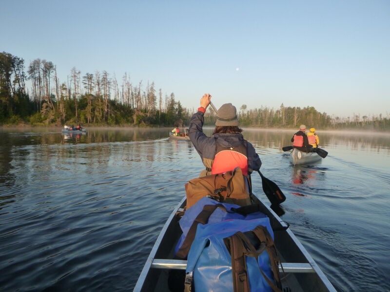 The image shows people canoeing on a calm lake. The person in the foreground is raising their hand. There are other canoes in the distance, and the water has a slight mist. The sky is clear, and the sun is likely rising or setting.